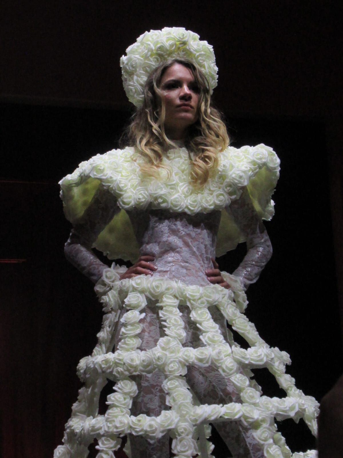 Model in a runway dress made of white roses, hat, and architectural skirt, hands on hips.