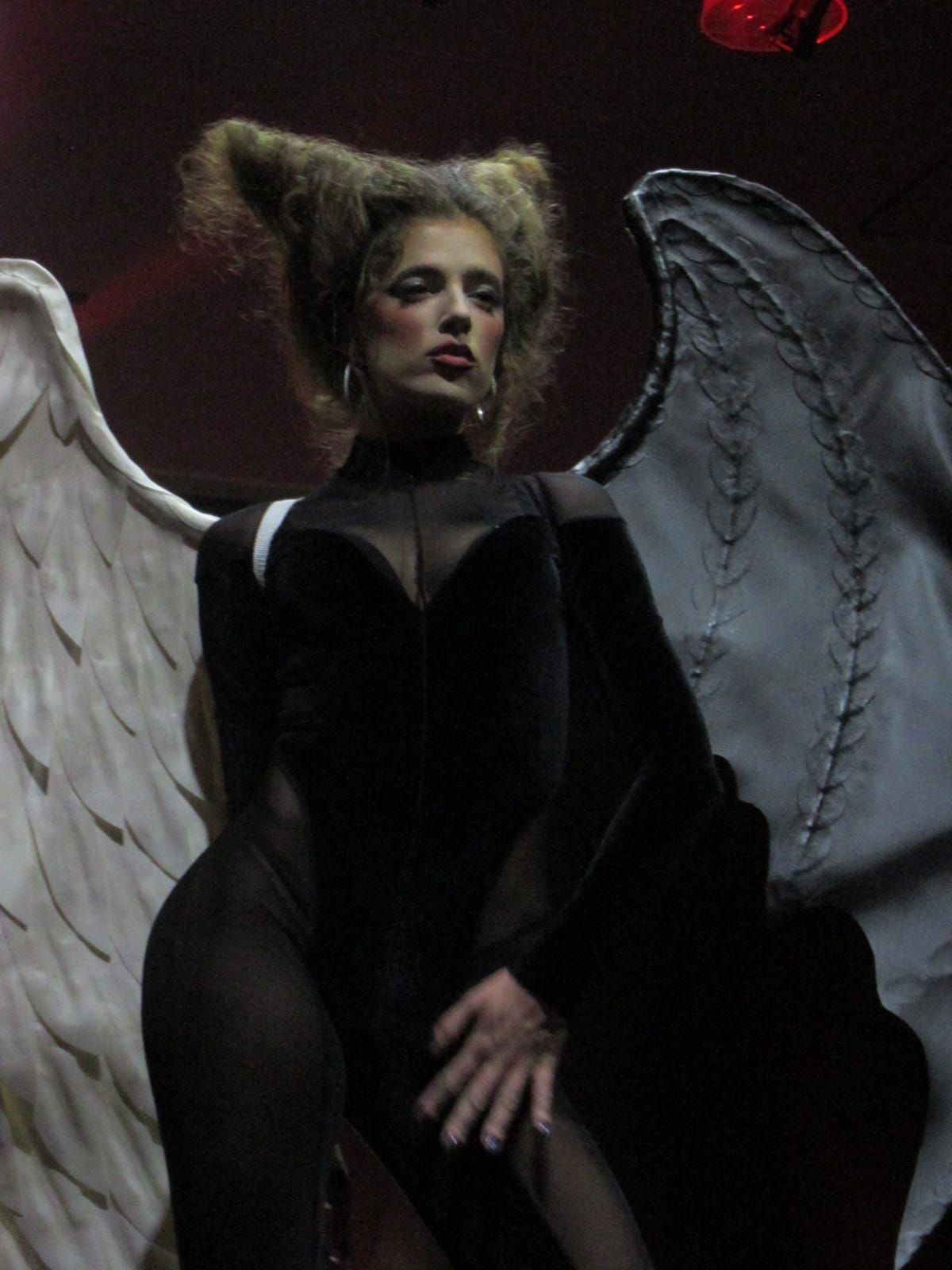 Woman in black devil/angel costume, wings, theatrical makeup, red background.