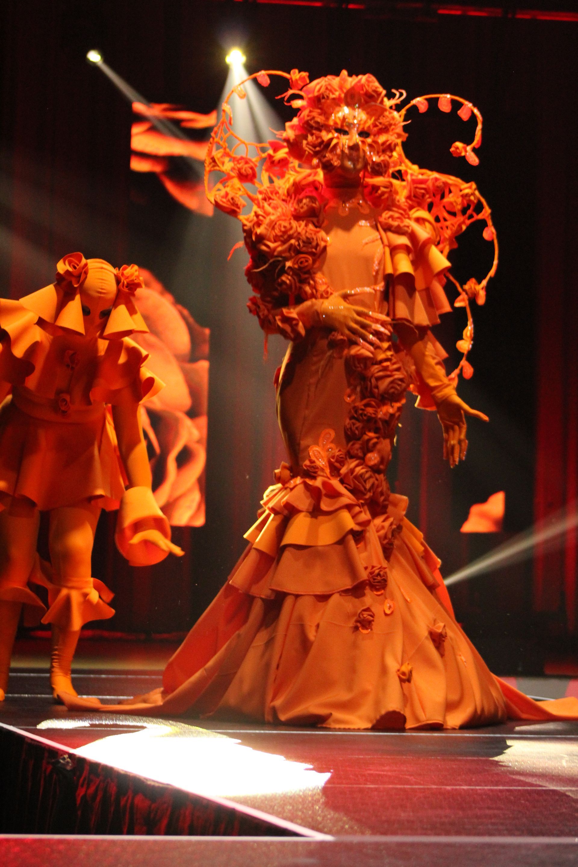 A performer in a flowing orange gown and elaborate headdress on stage, spotlighted.