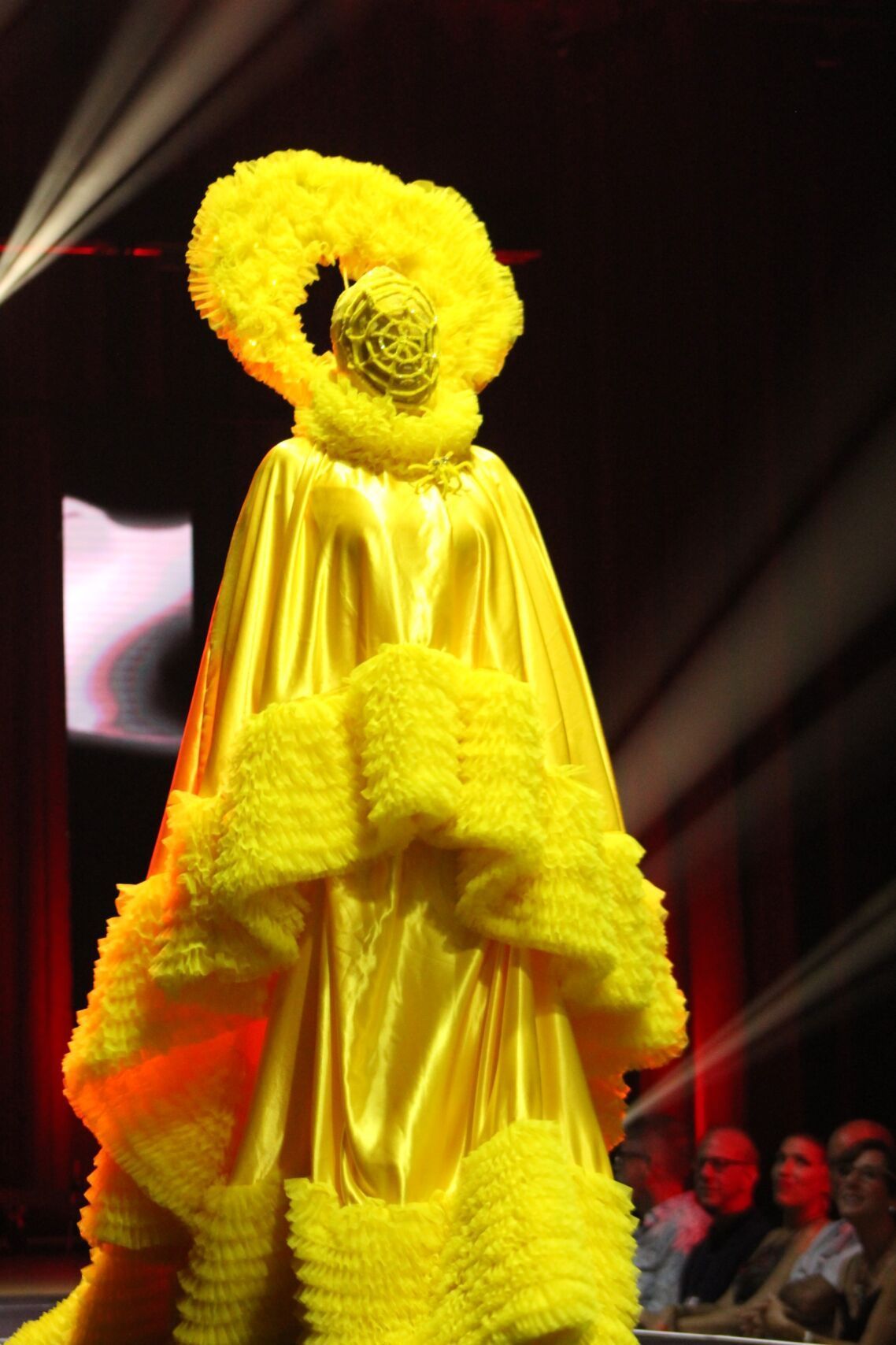 Woman in bright yellow runway fashion: ornate headpiece, ruffled detailing, satin fabric.