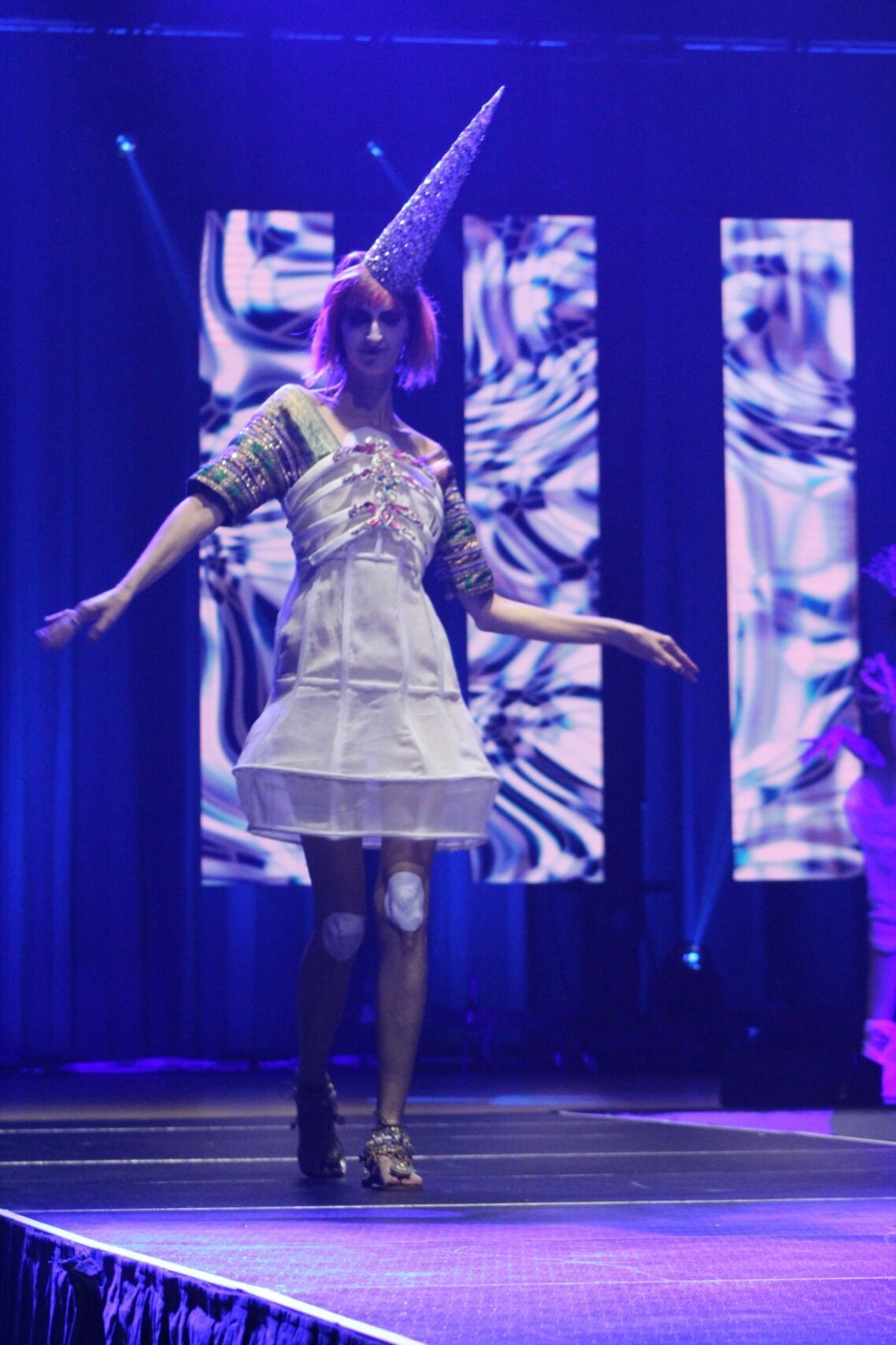 Woman on runway wearing a whimsical outfit: cone headpiece, embellished bodice, knee decorations. Stage backdrop with blue light.