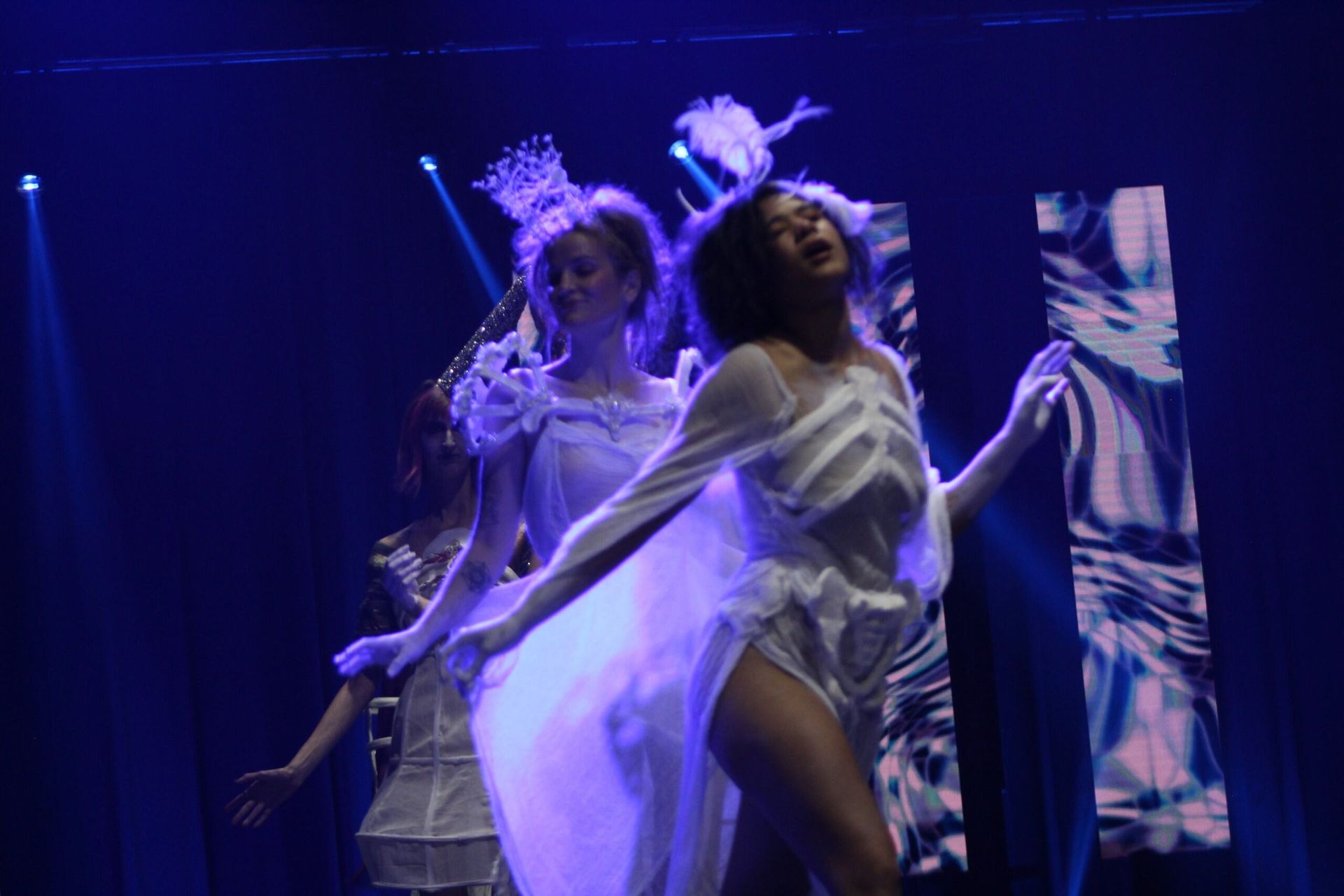 Dancers in white costumes on stage with blue lighting, arms outstretched.