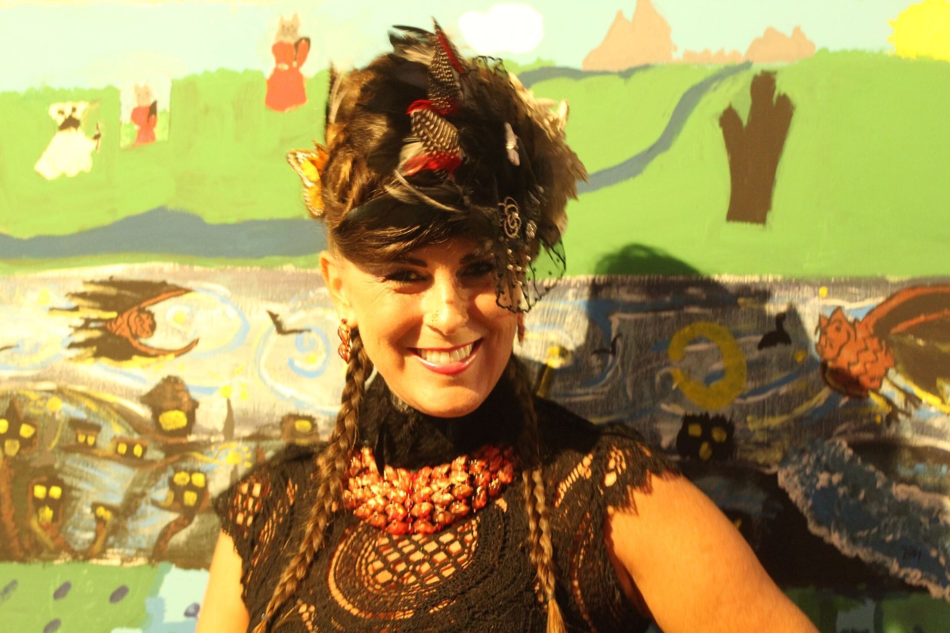 Woman with elaborate feather headpiece, smiling in front of a colorful mural.