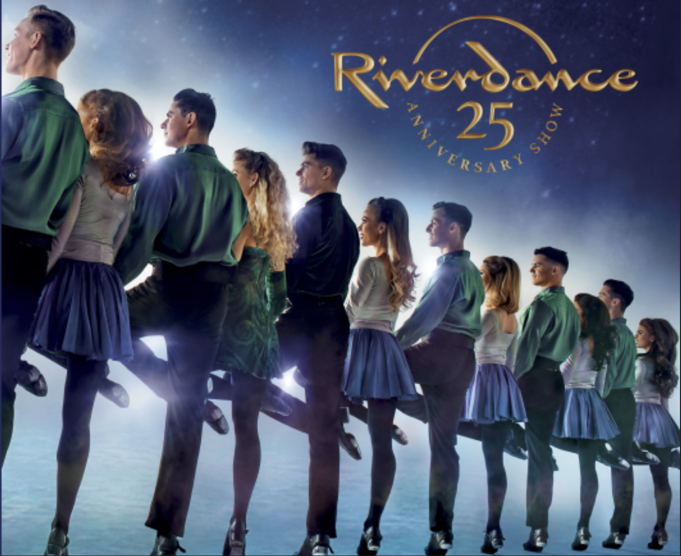 Riverdance dancers in a line, performing on a stage. Performers wear blue skirts, green/blue shirts. 