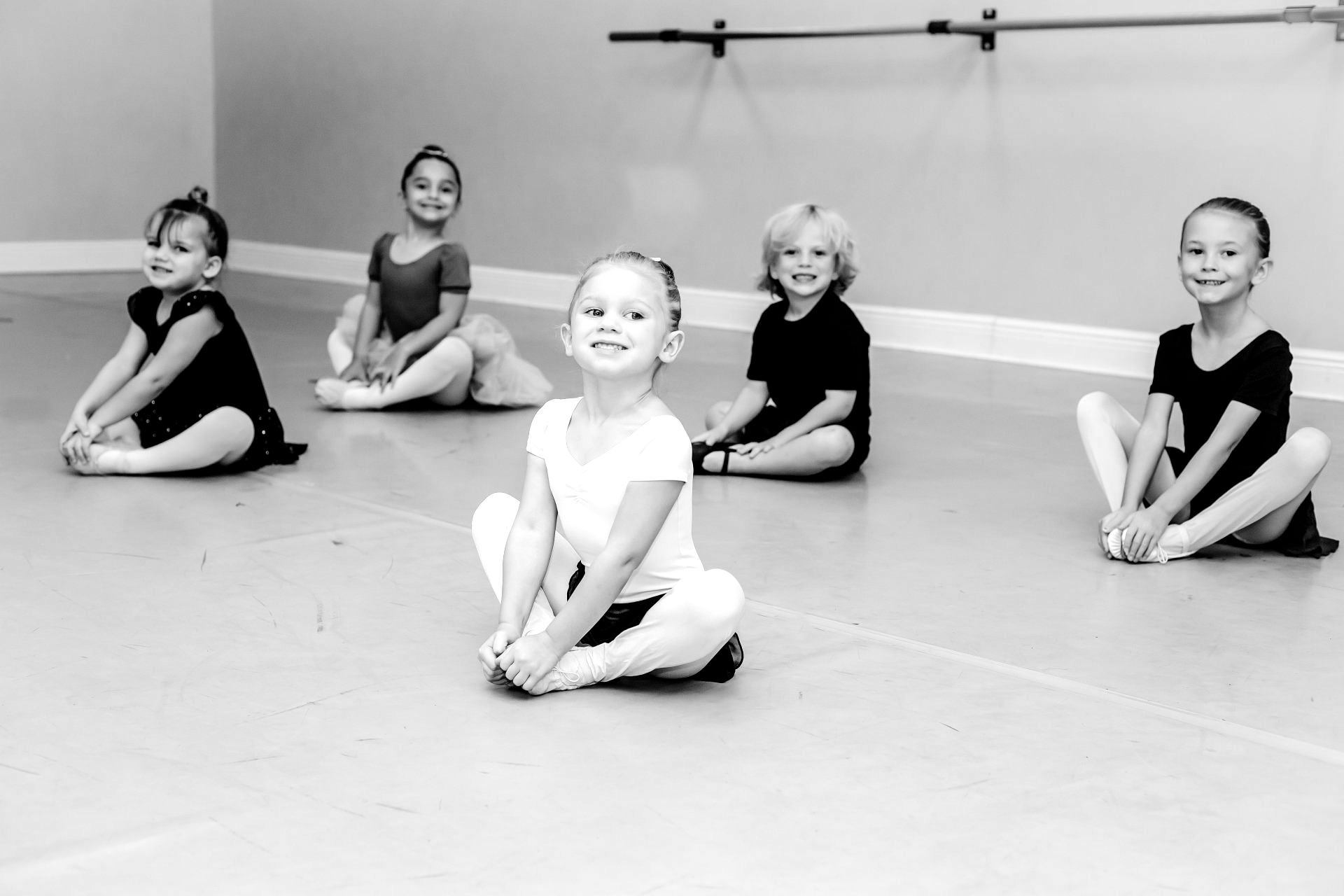 Five young dancers in a studio, sitting cross-legged and smiling. Black and white.