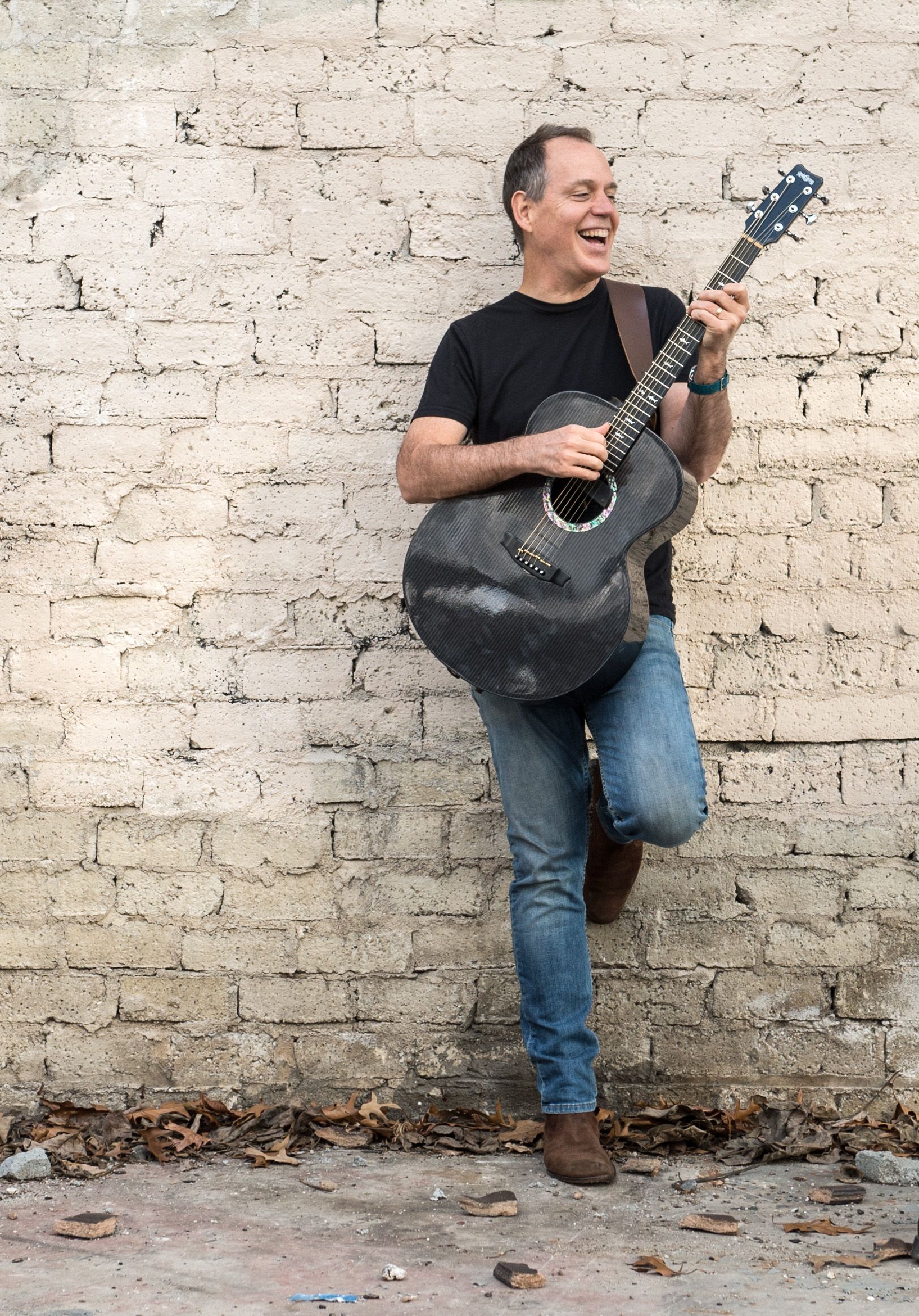 Man leaning against a brick wall, smiling and playing a blue guitar.