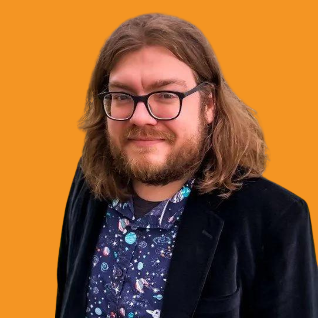 A man with long brown hair and glasses smiles. He wears a space-themed shirt and a dark jacket against an orange backdrop.
