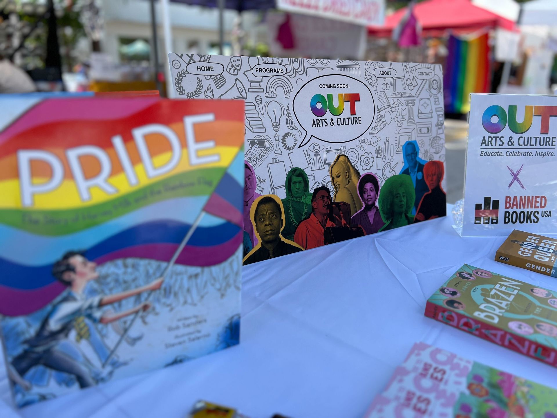 A display of LGBTQ+ books at an outdoor event. Colorful Pride flag and 