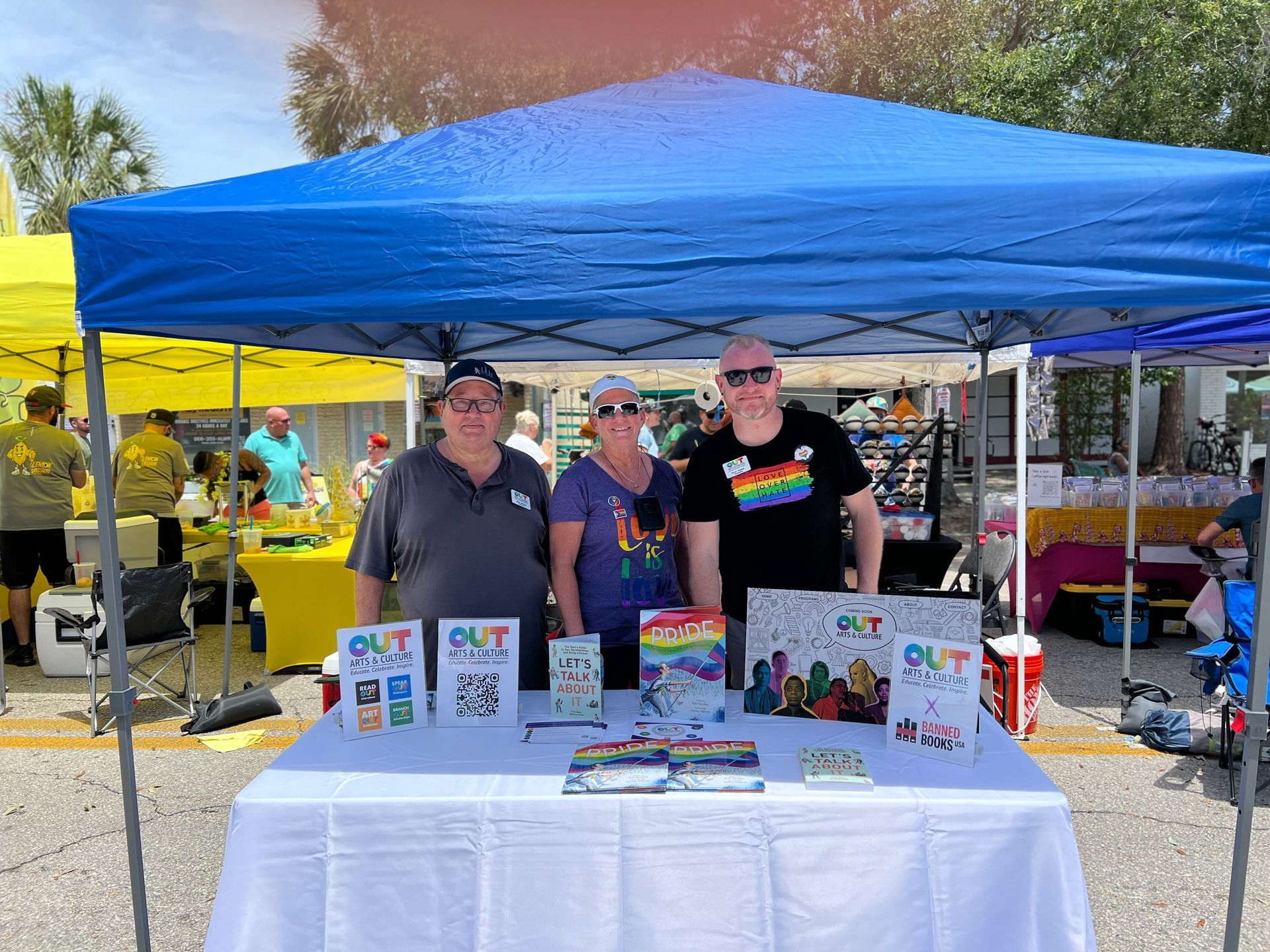 Three people at a Pride booth under a blue tent, displaying 