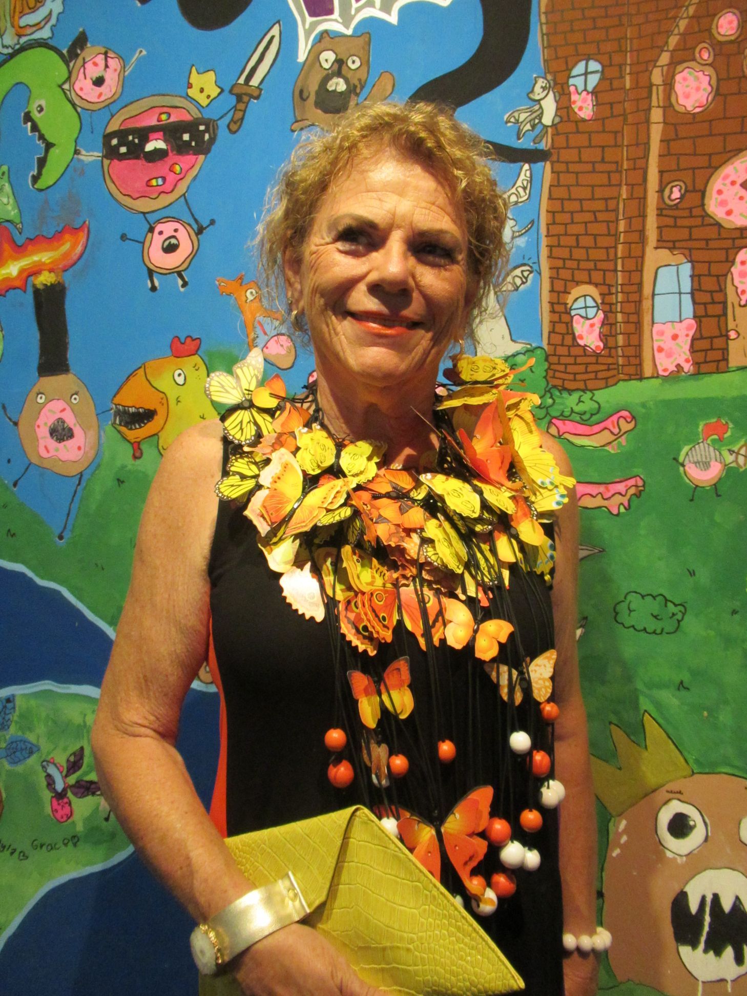 Woman in a black dress with a colorful leaf-like necklace and clutch, in front of a whimsical mural.