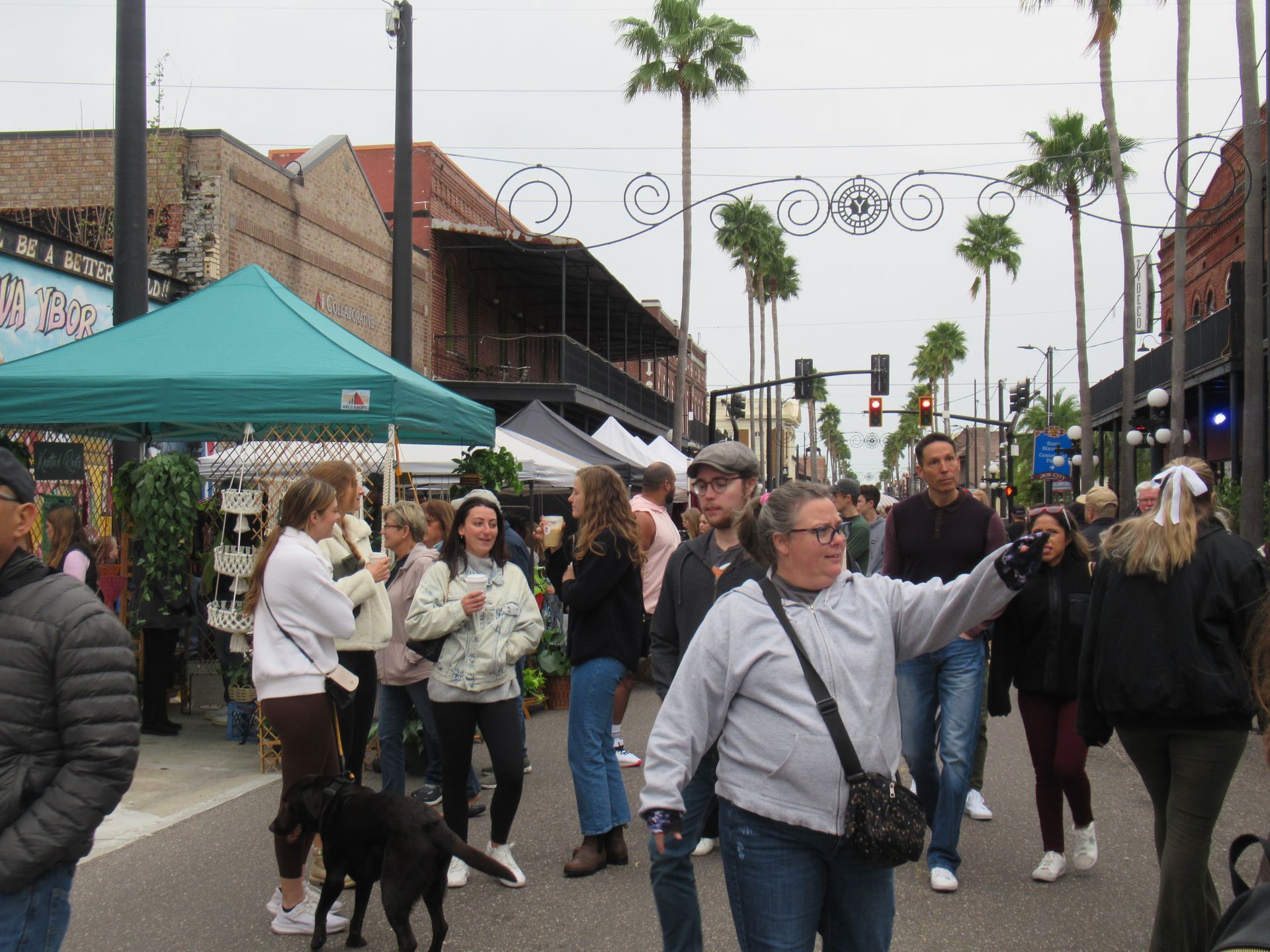 Crowd at an outdoor market on a street lined with palm trees; people browsing, shopping, some smiling.