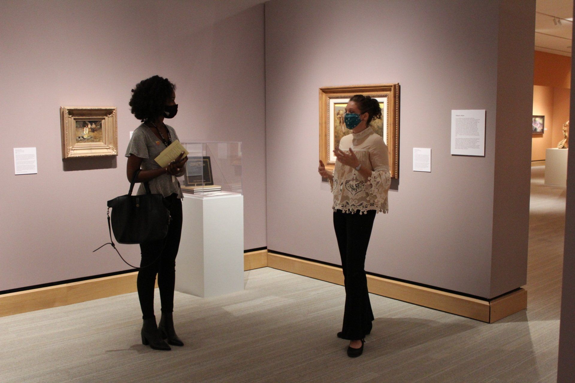 Two women wearing masks in an art gallery, looking at a painting. Soft mauve walls, tan trim.