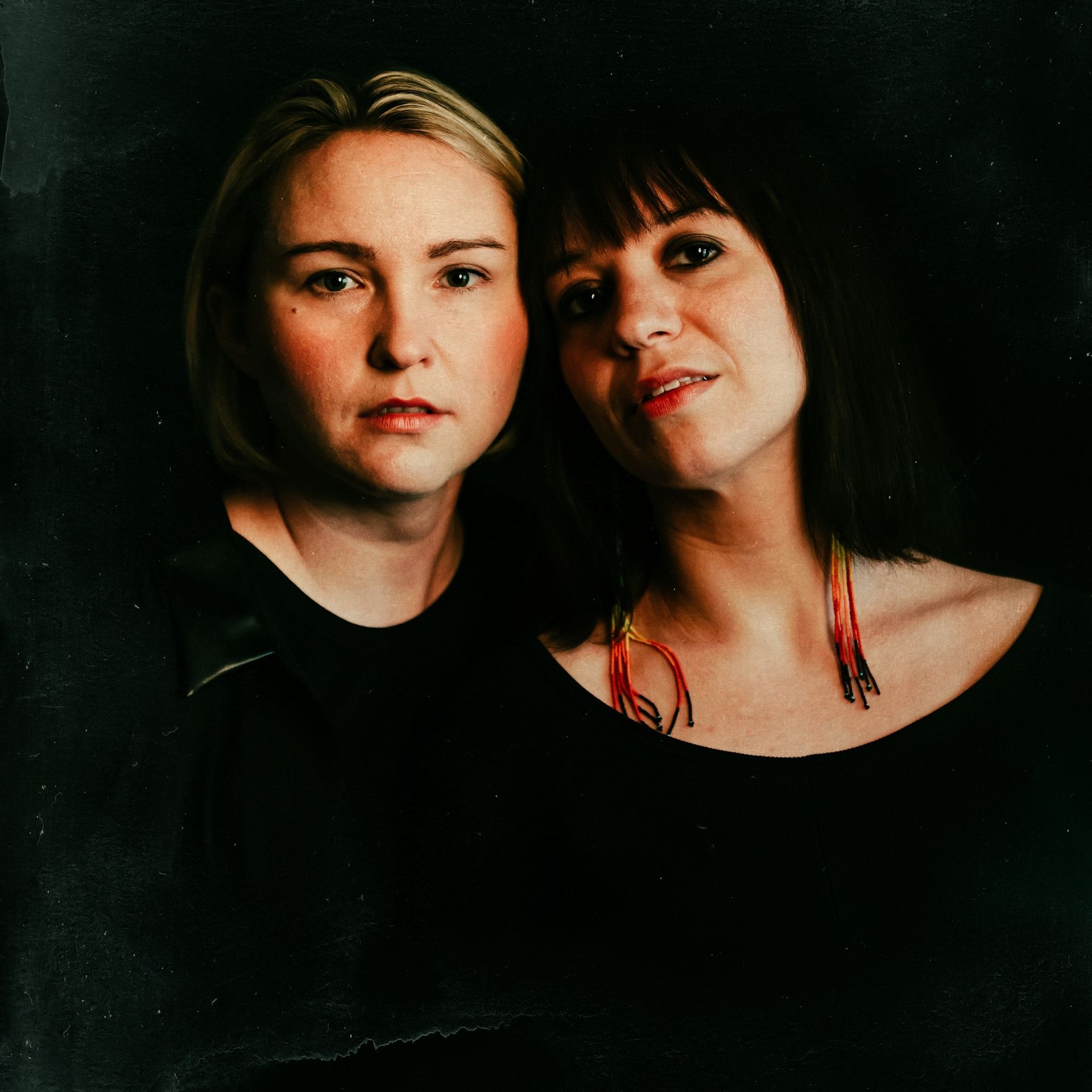 Two women, blonde and brunette, looking at the camera. Both wearing black tops, dark background.