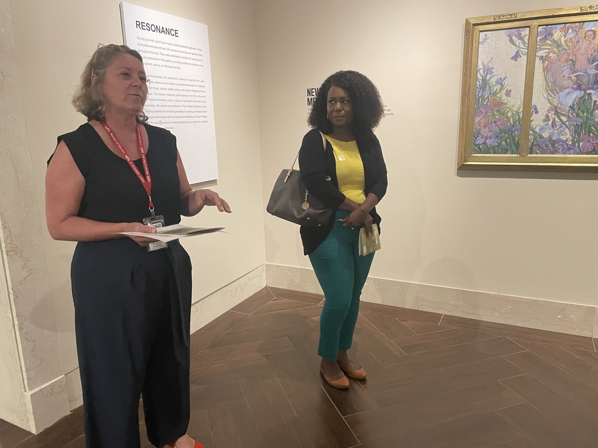 Woman giving tour in gallery, explaining art to another woman standing by a painting.
