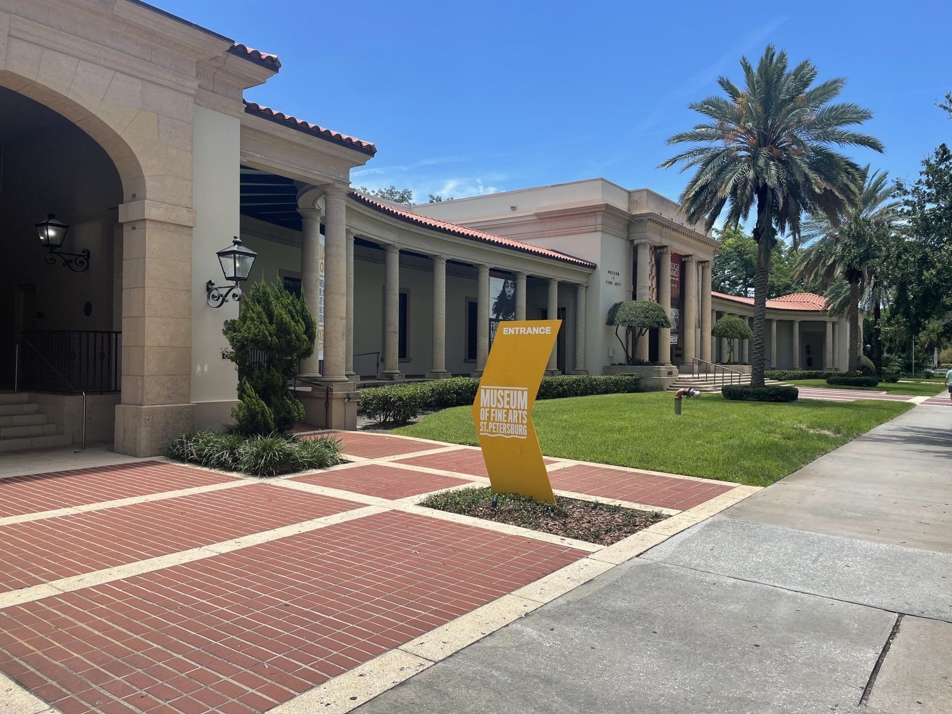 A yellow sign in front of a Mediterranean-style building with brick walkways and palm trees under a blue sky.