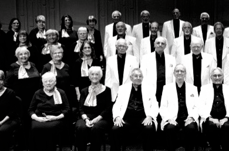 Choir of older adults in formal wear, posing for a group photo. Black and white.