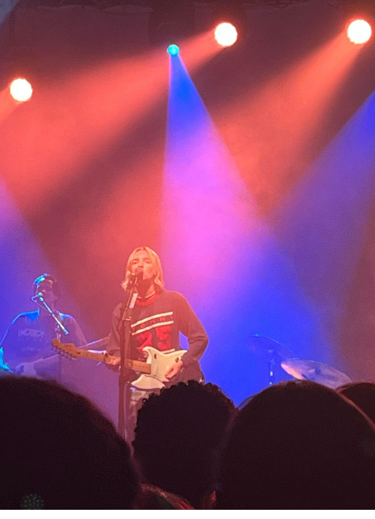 Singer on stage, playing guitar under blue and red lights, performing for a crowd.