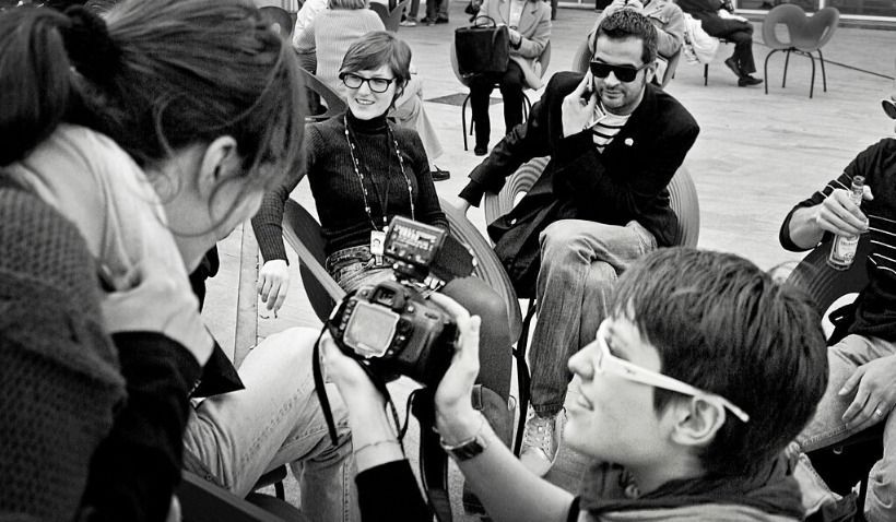 People in a group, some holding cameras, posing and taking pictures. Woman in glasses smiles.