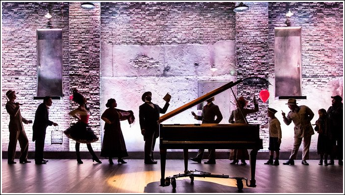 Silhouetted figures on stage around piano; brick backdrop, spotlights.