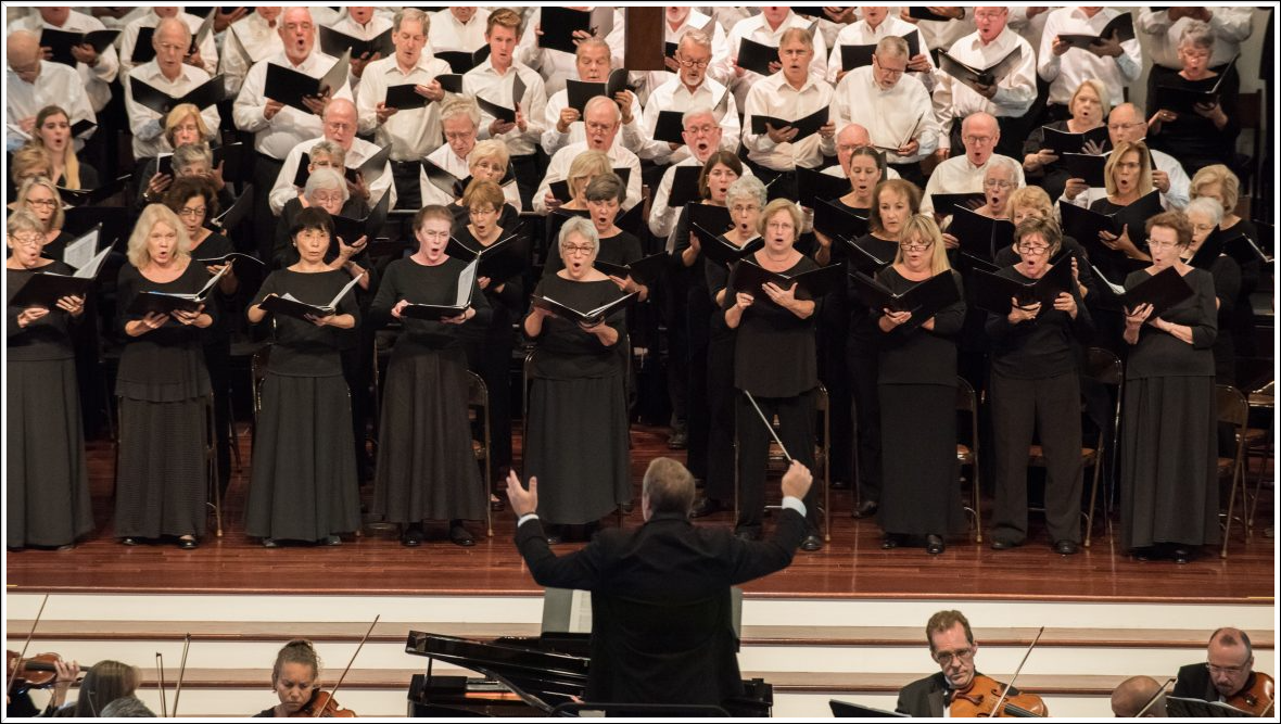 A choir and orchestra performing on stage; conductor facing forward.