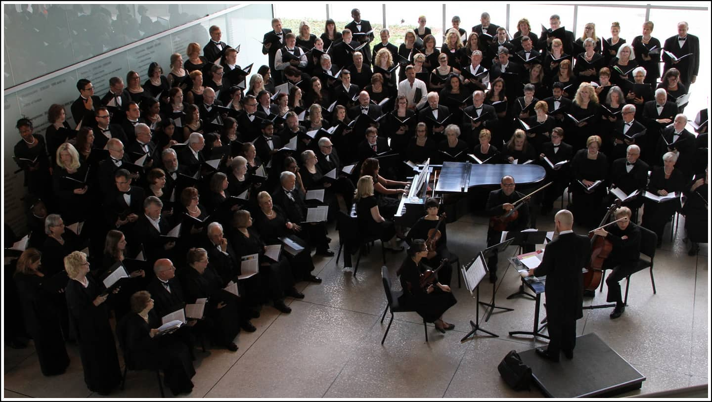 A large choir and orchestra in black attire performing on stage, with a conductor facing them.
