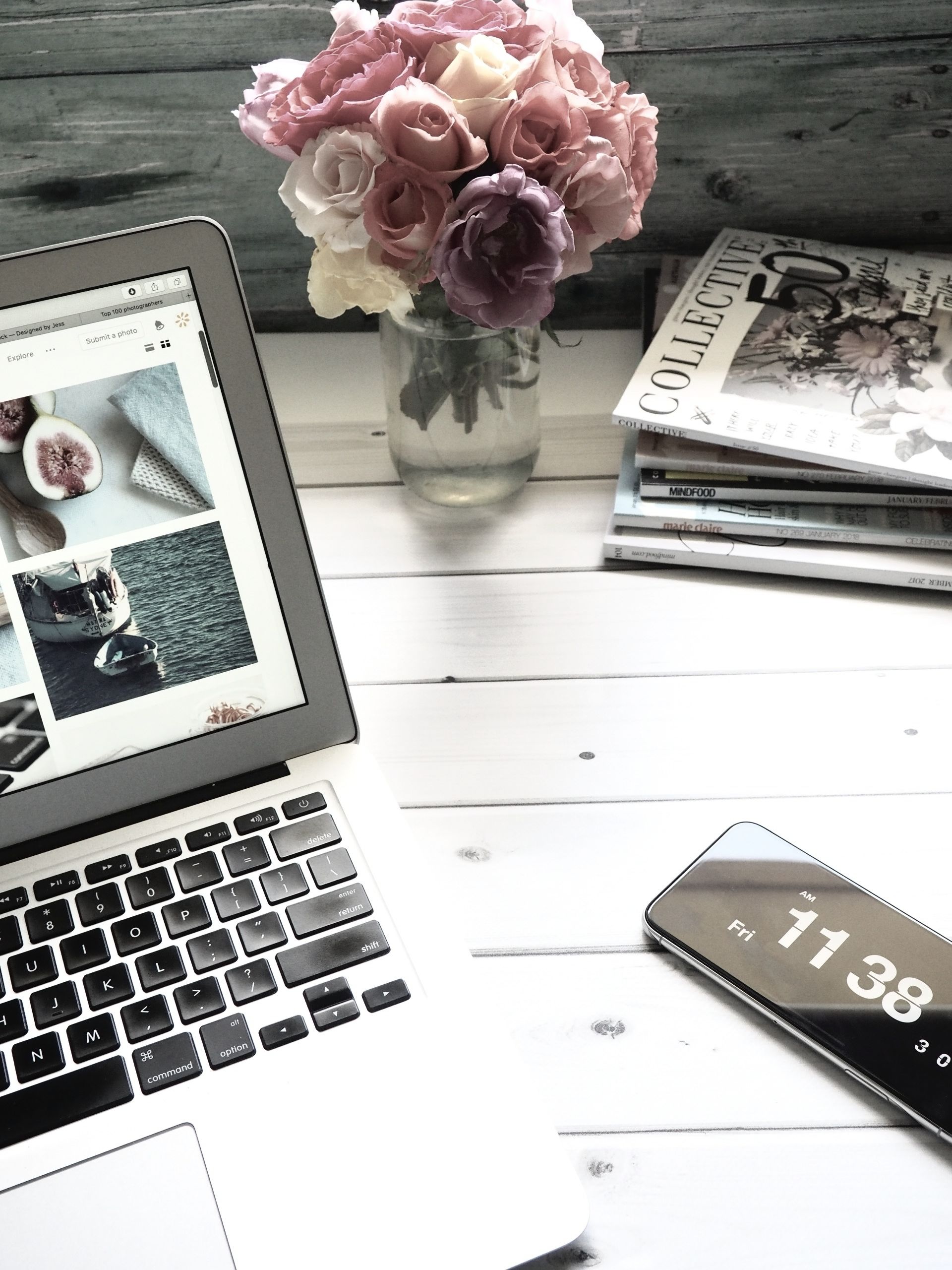 Laptop on desk, next to flowers and magazines, with a phone showing the time.