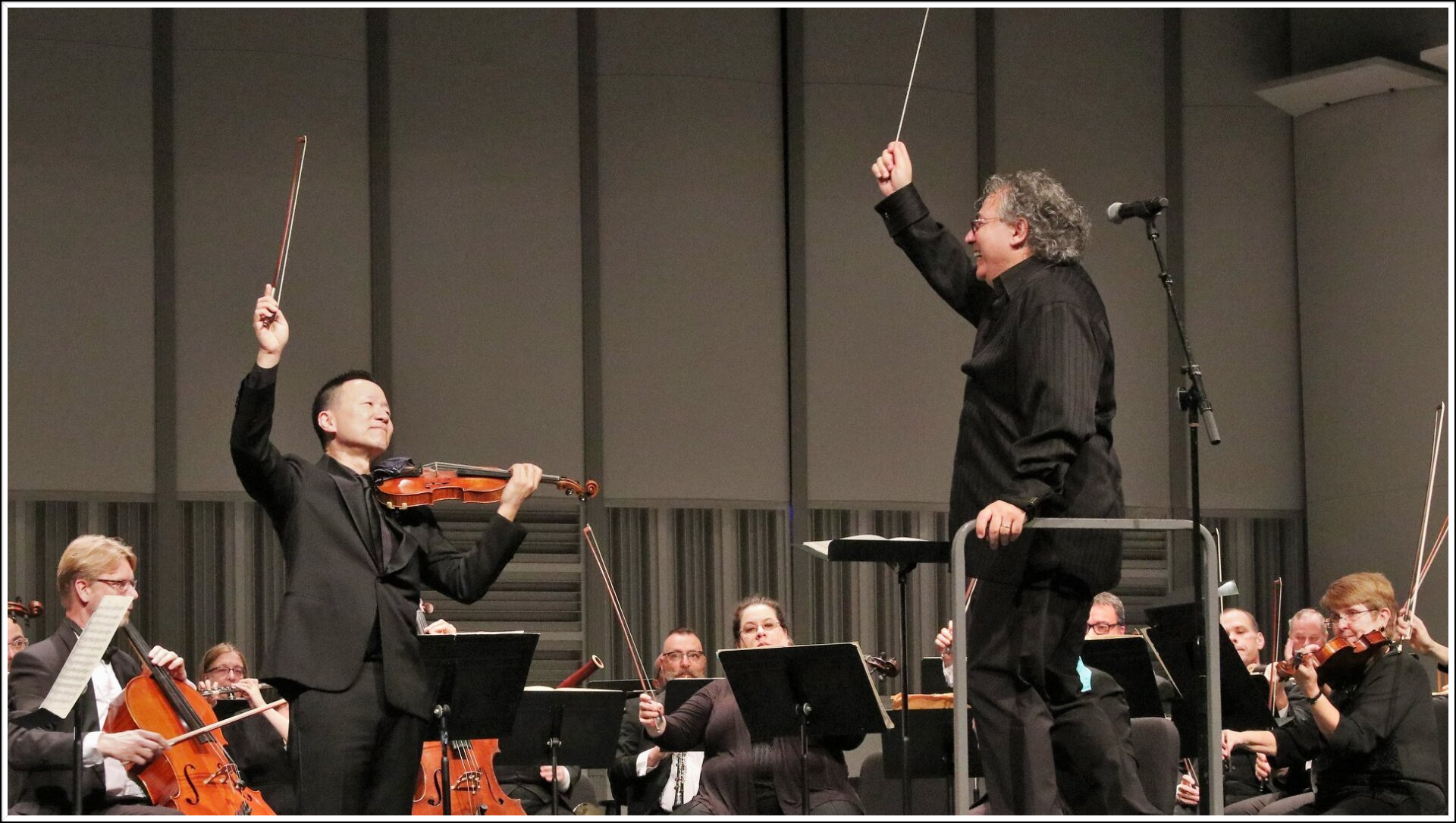 Violinist and conductor on stage, both holding bows in air. Orchestra behind them in concert hall.