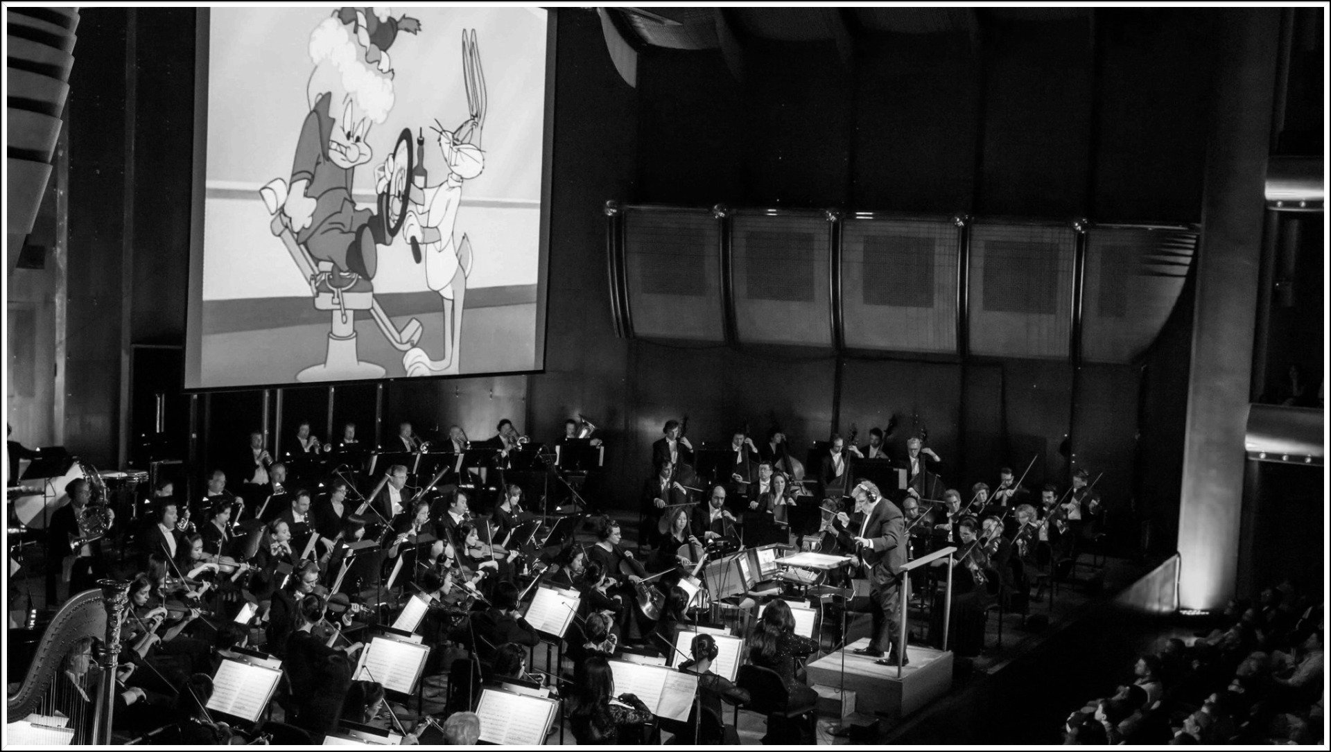 Black and white photo of orchestra playing live music with cartoon on the big screen.
