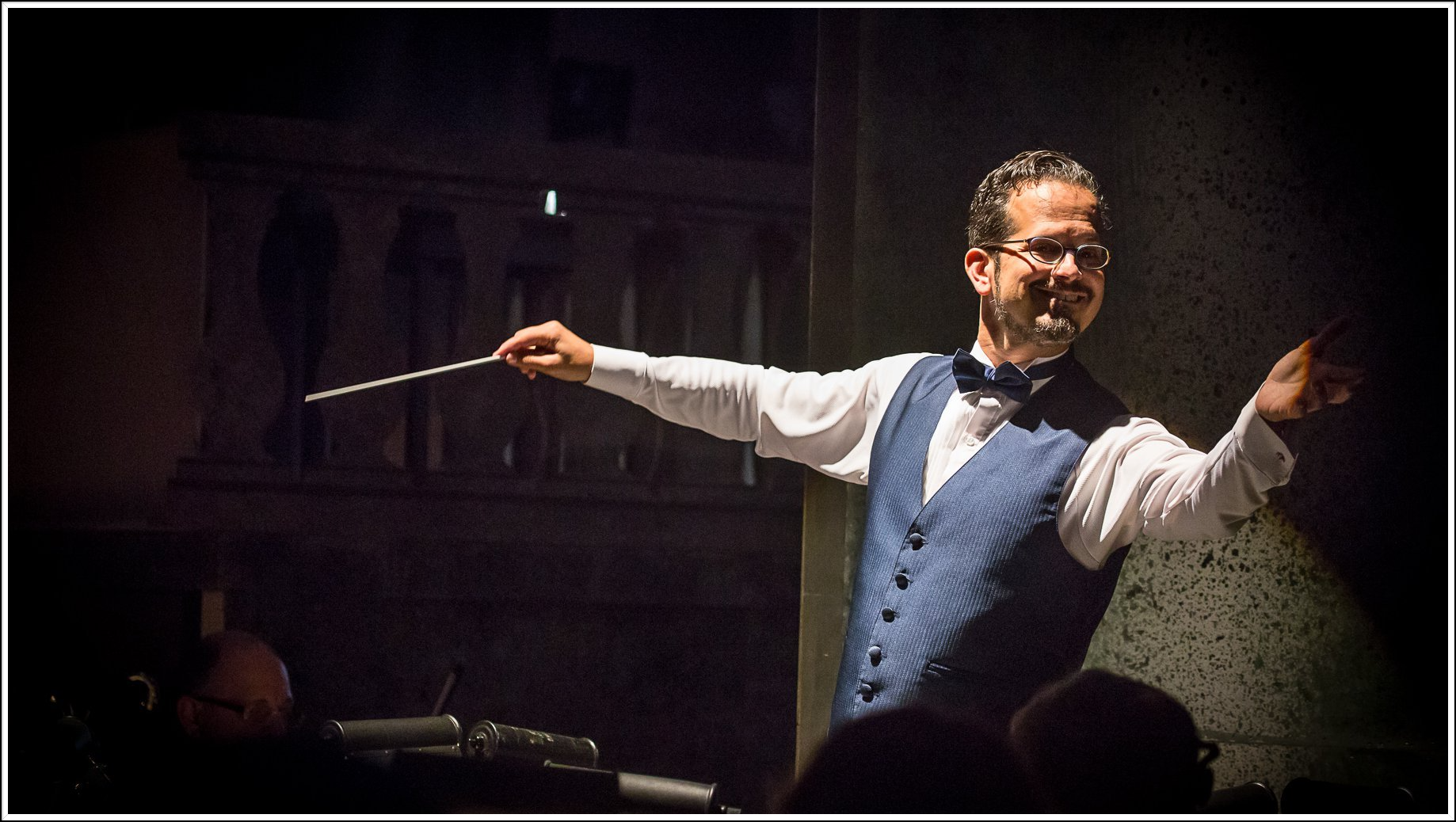 A conductor with a baton directs an orchestra, wearing a blue vest and smiling, in a dark hall.
