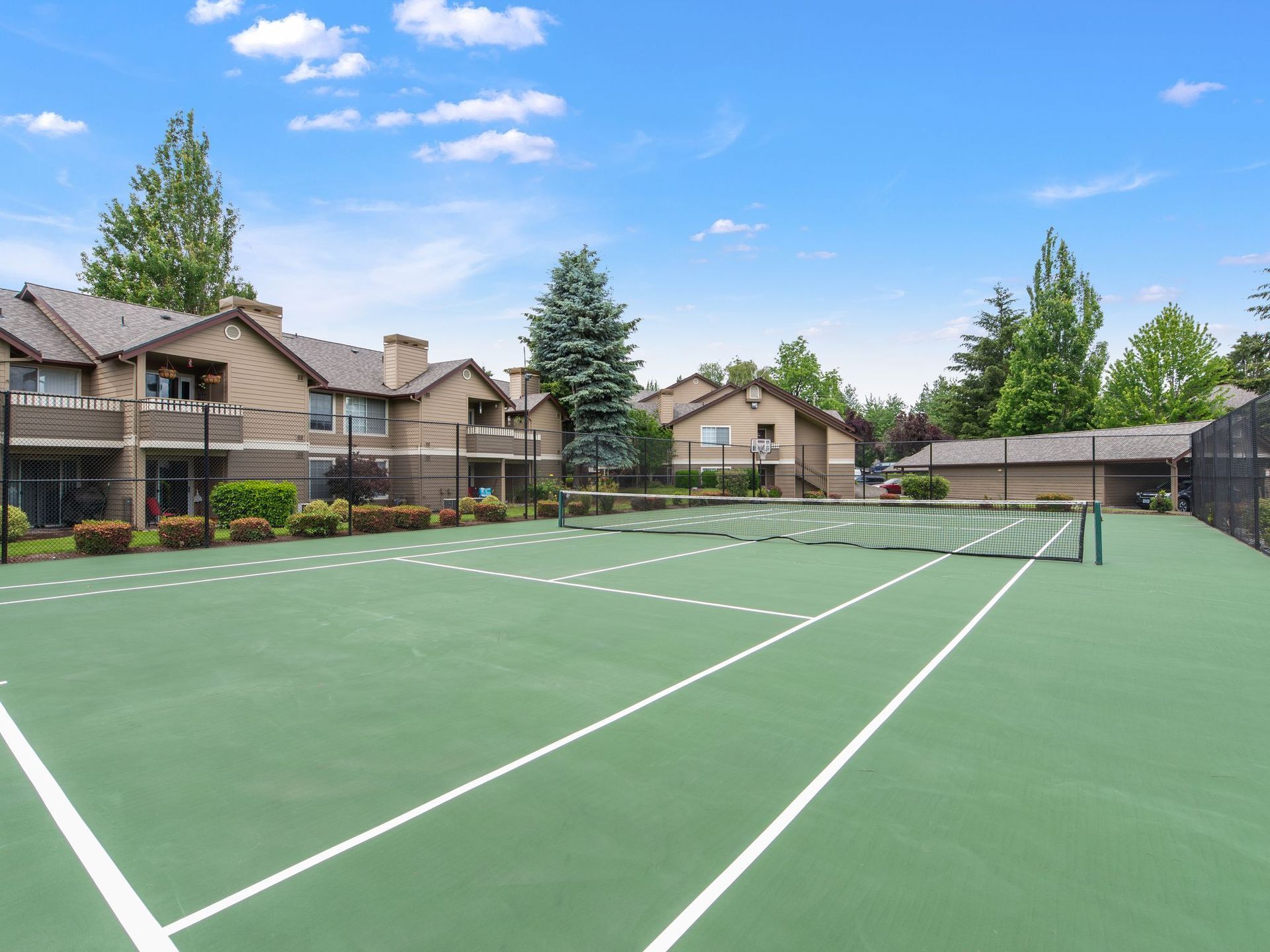 Outdoor tennis court with green surface beside townhouses under a clear blue sky