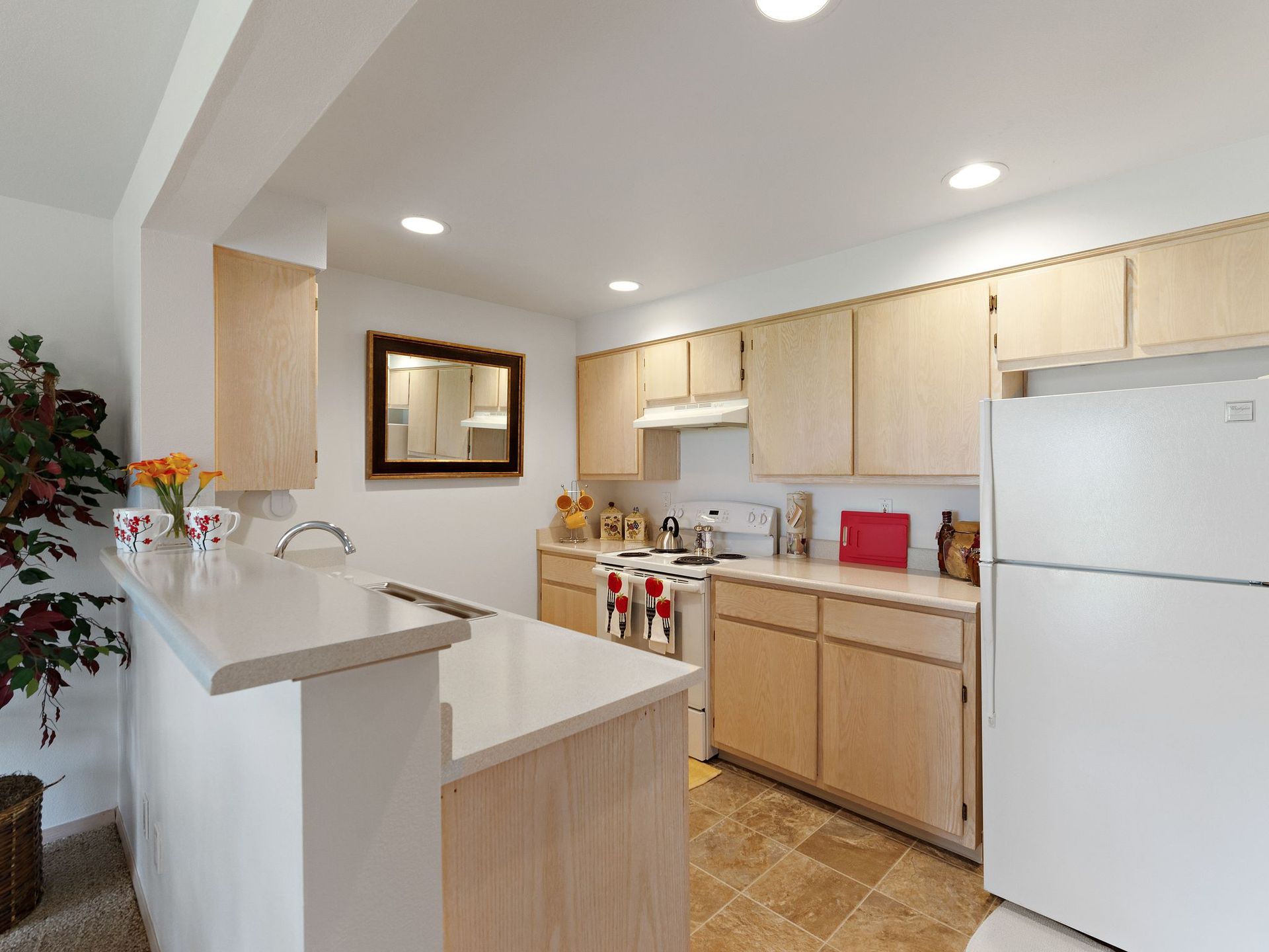 Bright kitchen with white appliances, light wood cabinets, tan tile floor, and a breakfast counter