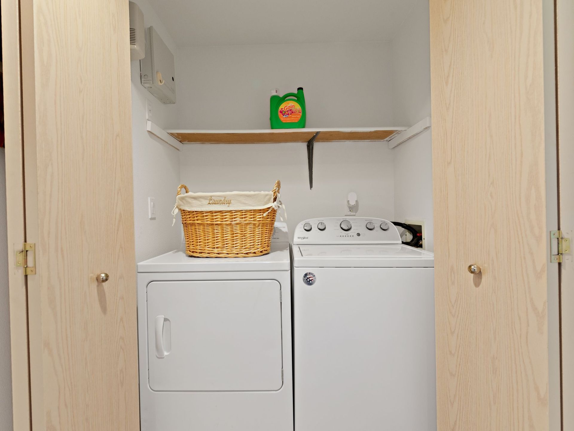 Laundry closet with a washer, dryer, wicker basket, and shelf holding a green container