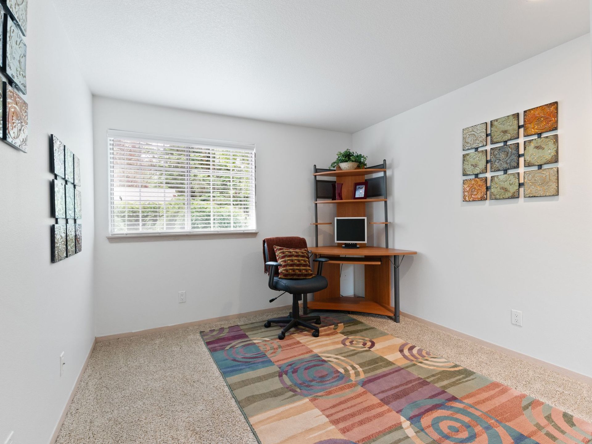 Small home office with a desk, chair, window, wall art, and colorful rug in a bright room