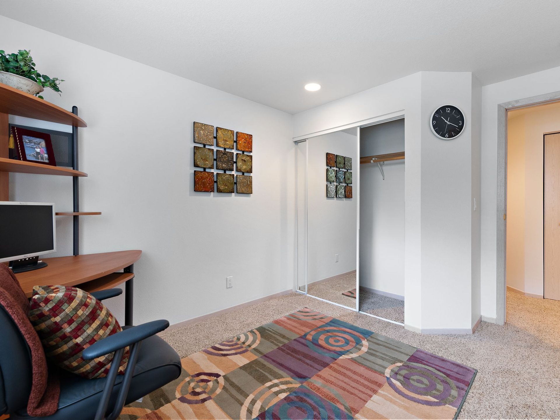 Small home office with desk, chair, colorful rug, wall art, and open closet door.