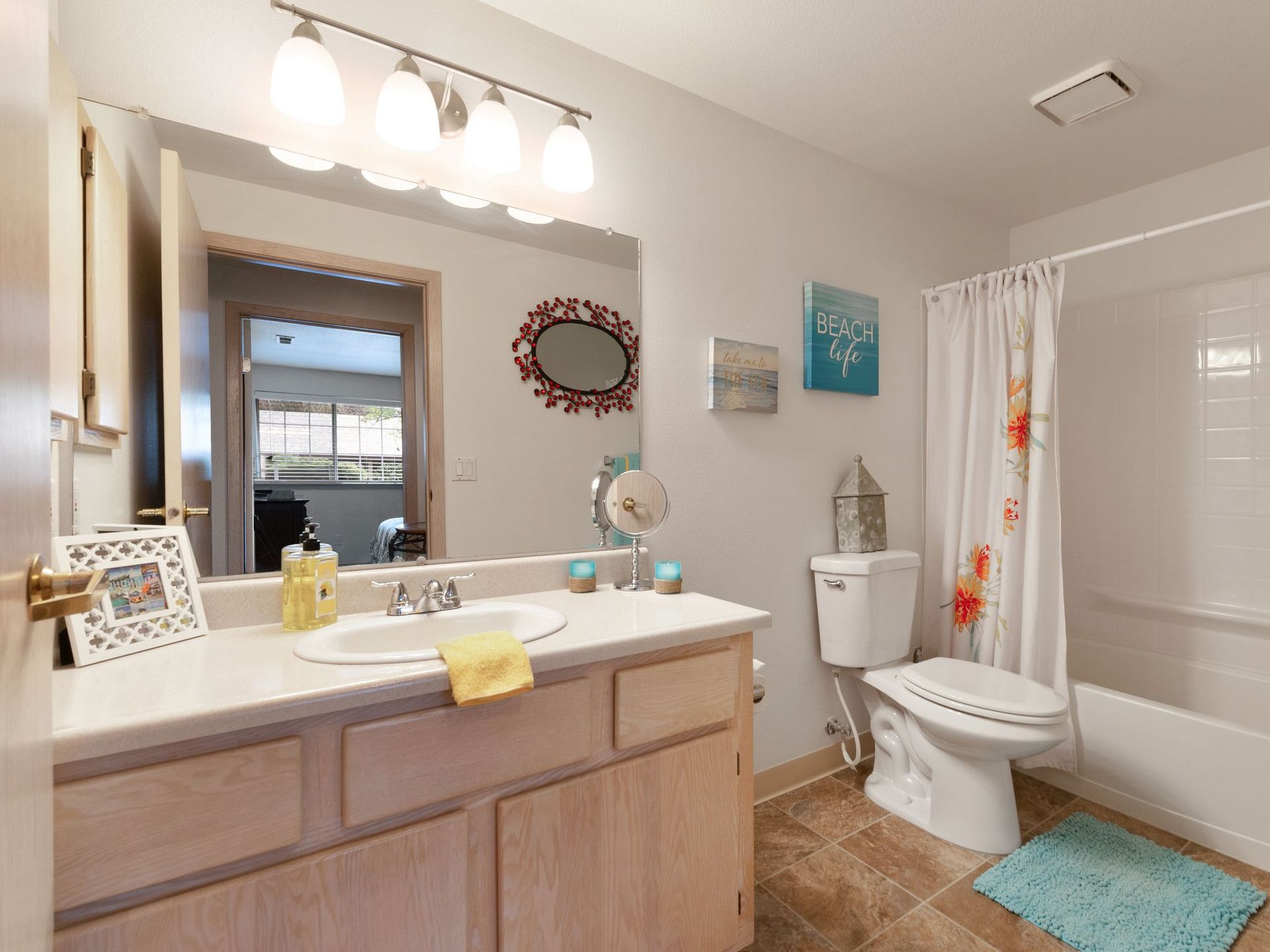 Bright bathroom with wood vanity, white toilet, tub, teal rug, and decorative wall art under three lights
