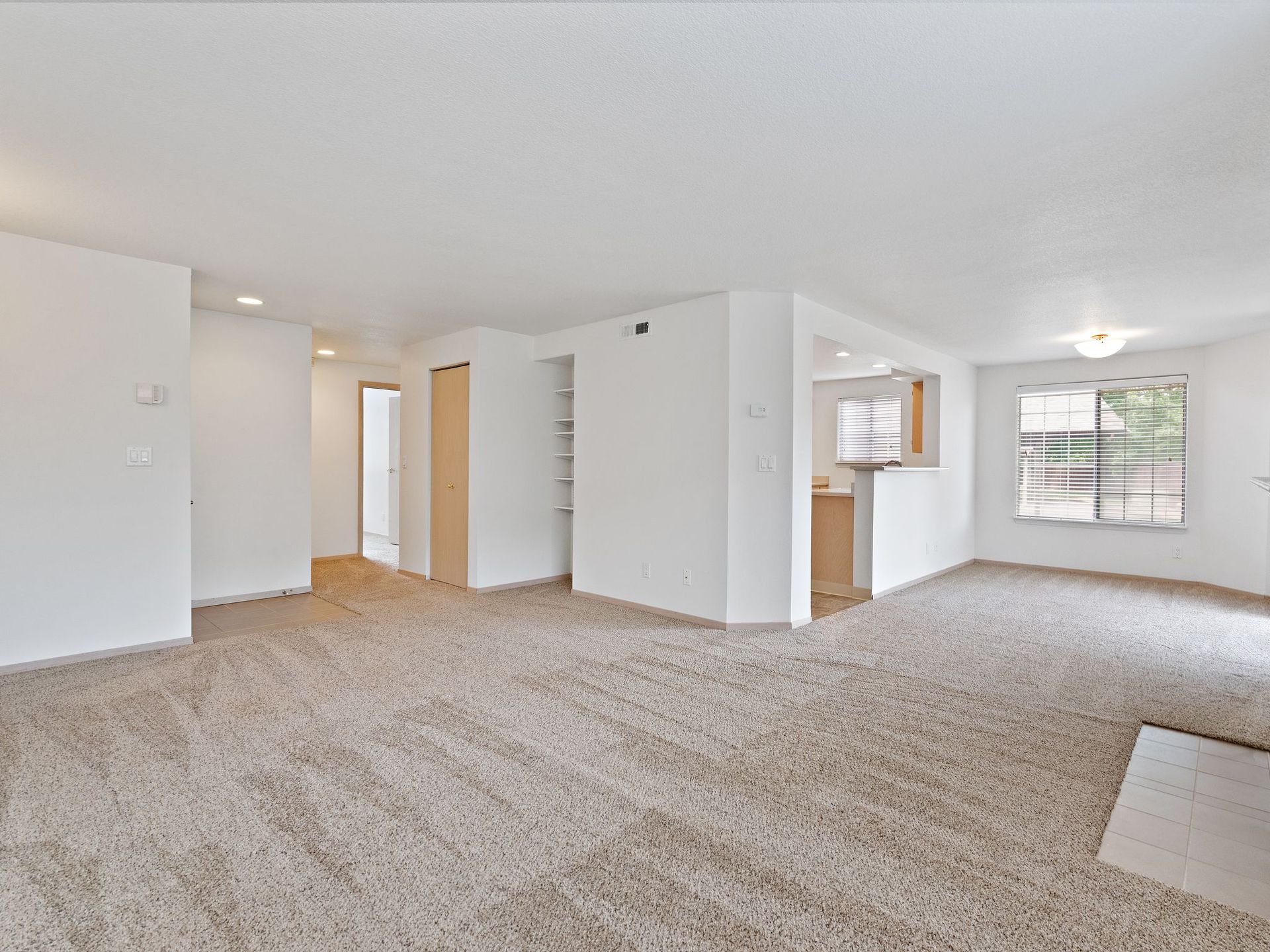 Empty carpeted living room with white walls, open kitchen pass-through, and large window with blinds