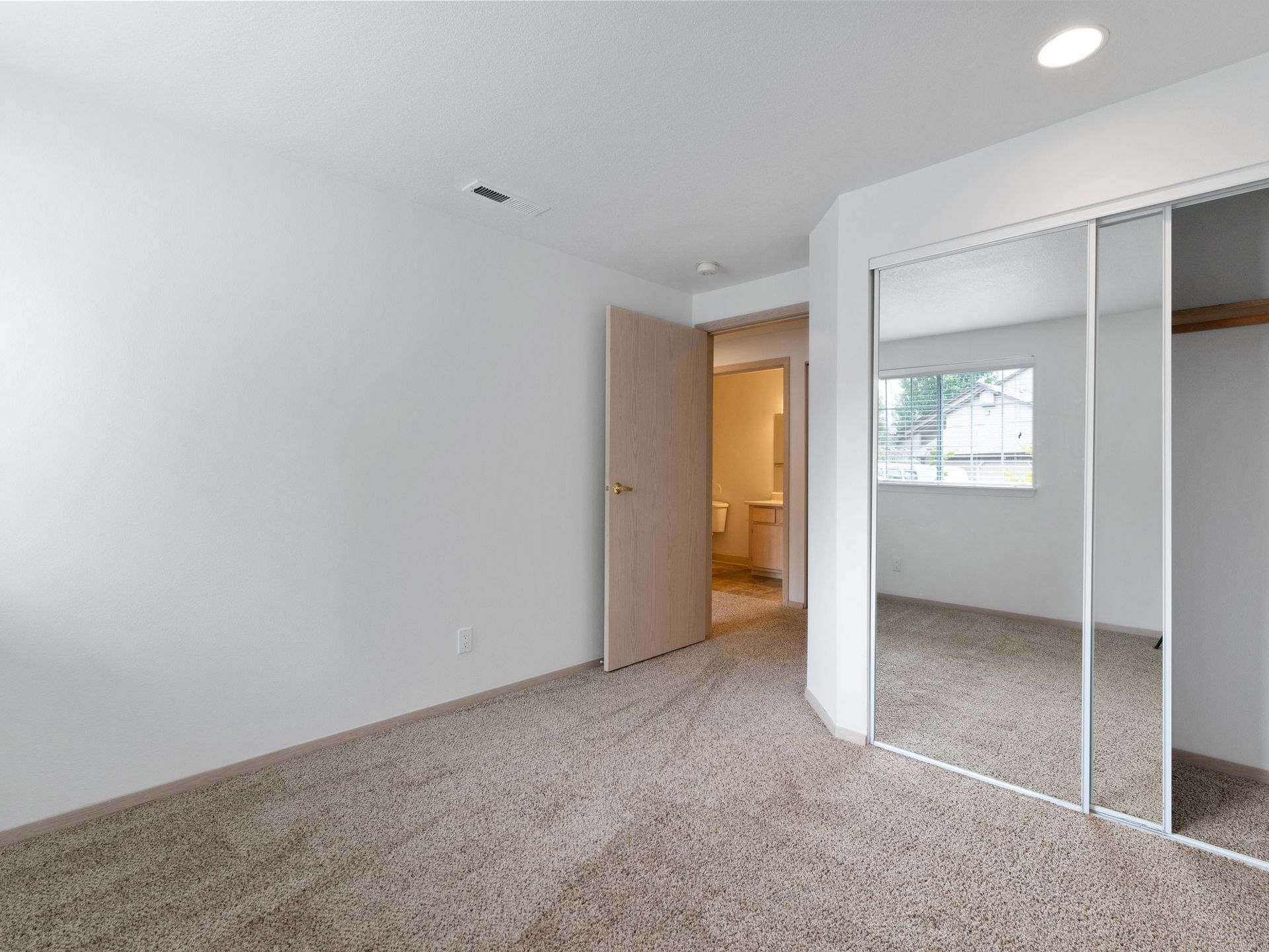 Empty beige bedroom with carpet, white walls, mirrored closet doors, and an open door to a lit hallway