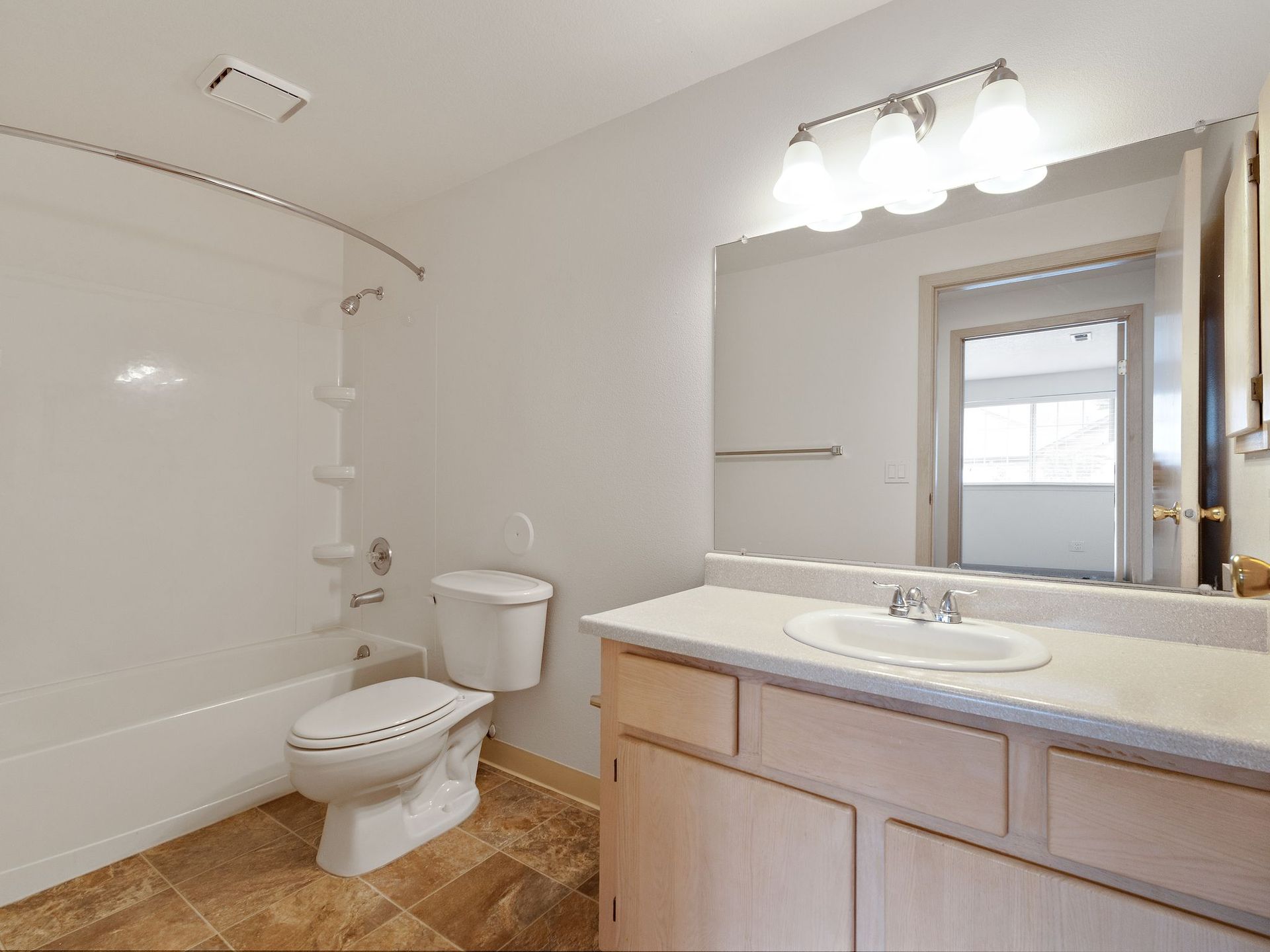 Bright bathroom with white tub, toilet, vanity, large mirror, and natural light from a window.