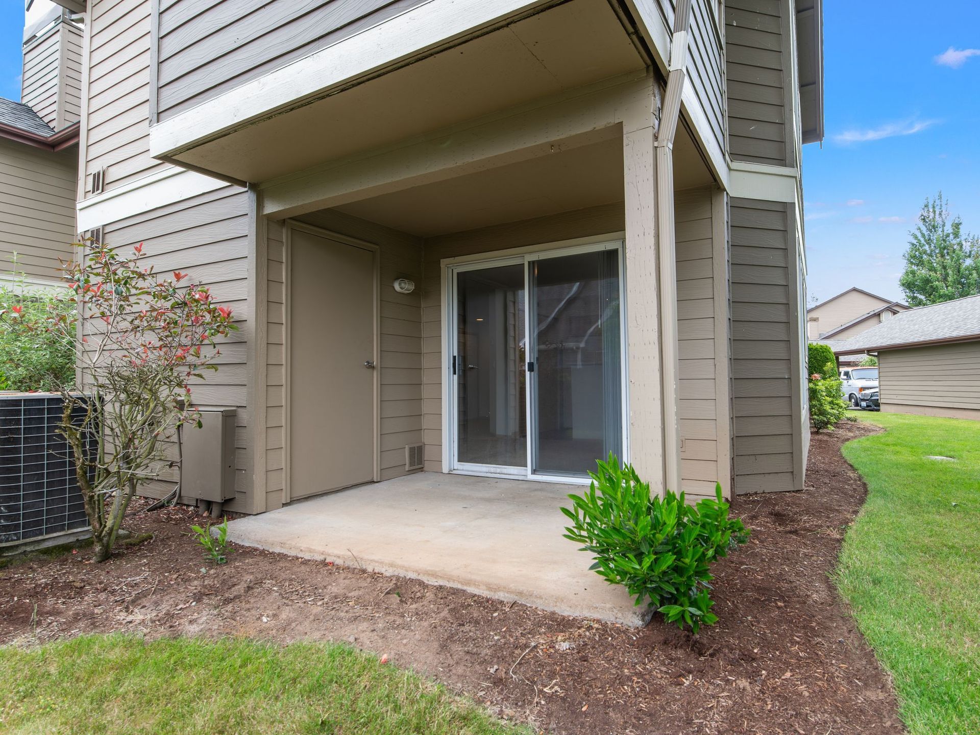 Patio entrance with sliding glass door, covered porch, tan siding, and landscaped yard.