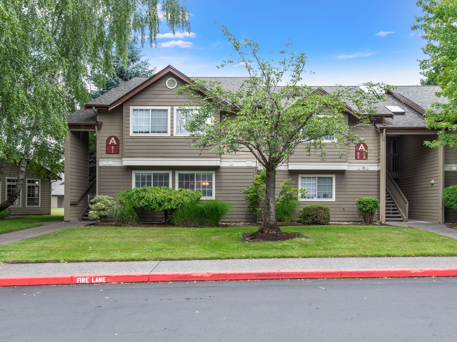 Two-story apartment building with beige siding, trees, shrubs, and a red curb in front