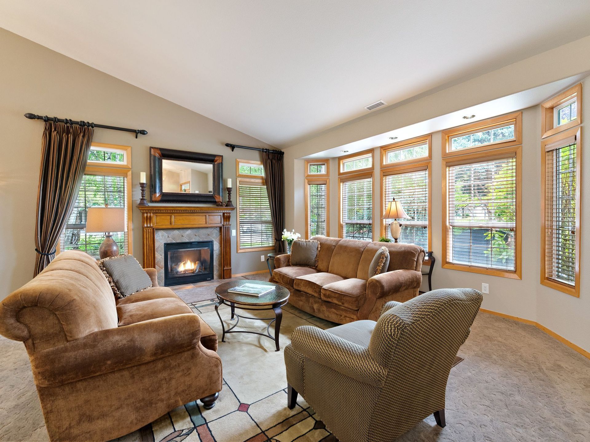 Cozy living room with fireplace, tan sofas, glass coffee table, and large bay windows with wood trim
