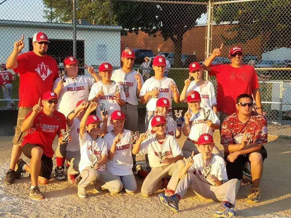 Baseball team posing in red and white uniforms on a field, with a chain-link fence and trees behind them.