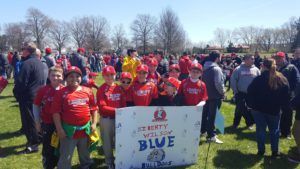 Crowd of people in red shirts gathered outdoors around a sign for the Blue event