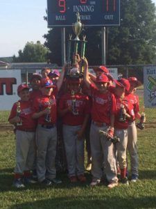 Youth baseball team in red uniforms celebrating with a trophy on the field.