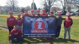 Youth baseball team in red jerseys posing outdoors behind a baseball banner on a grassy field.
