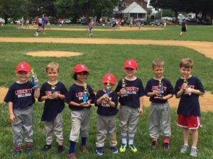 Seven young baseball players in navy shirts and red caps pose on a field, each holding a trophy.