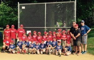 Youth baseball team in red jerseys posing with coaches in front of a chain-link backstop at a field