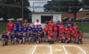 Youth baseball team posing for a group photo at home plate, in red and blue uniforms on a dirt field.