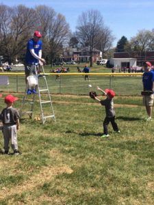 Kids playing baseball in a grassy park, with one child on a ladder holding a bat and others nearby.