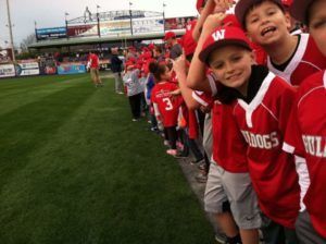 Young baseball fans in red jerseys line the field, smiling and waving beside the grass stadium aisle.
